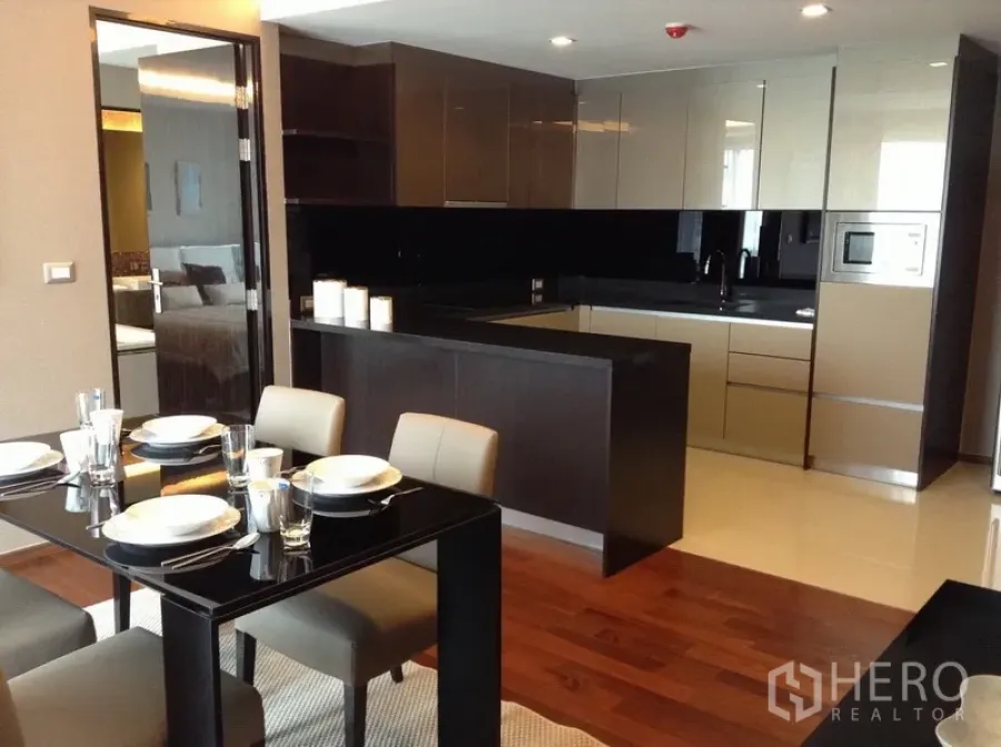Condominium for rent in Watthana, Bangkok - Modern dining area beside a U-shaped kitchen with glossy cabinets, black backsplash and wooden floors.