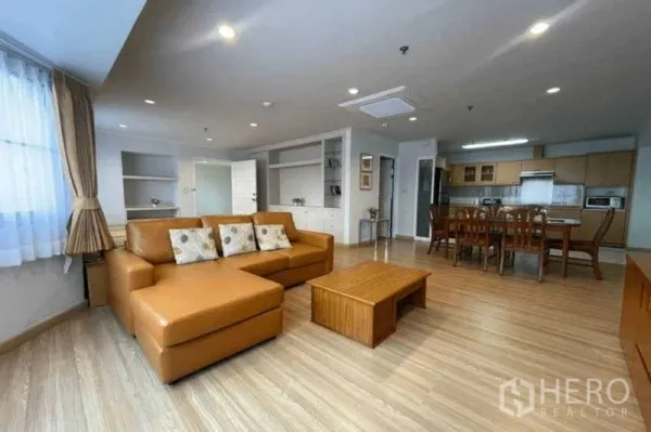 Condominium for rent in Watthana, Bangkok - Spacious lounge with L-shaped leather sofa, dining set and built-in shelves beside an open kitchen.