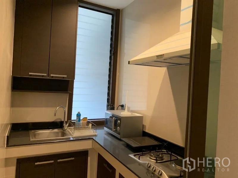 Townhouse for sale in Watthana, Bangkok - Compact enclosed kitchen with gas hob, range hood, sink and microwave near a tall window.