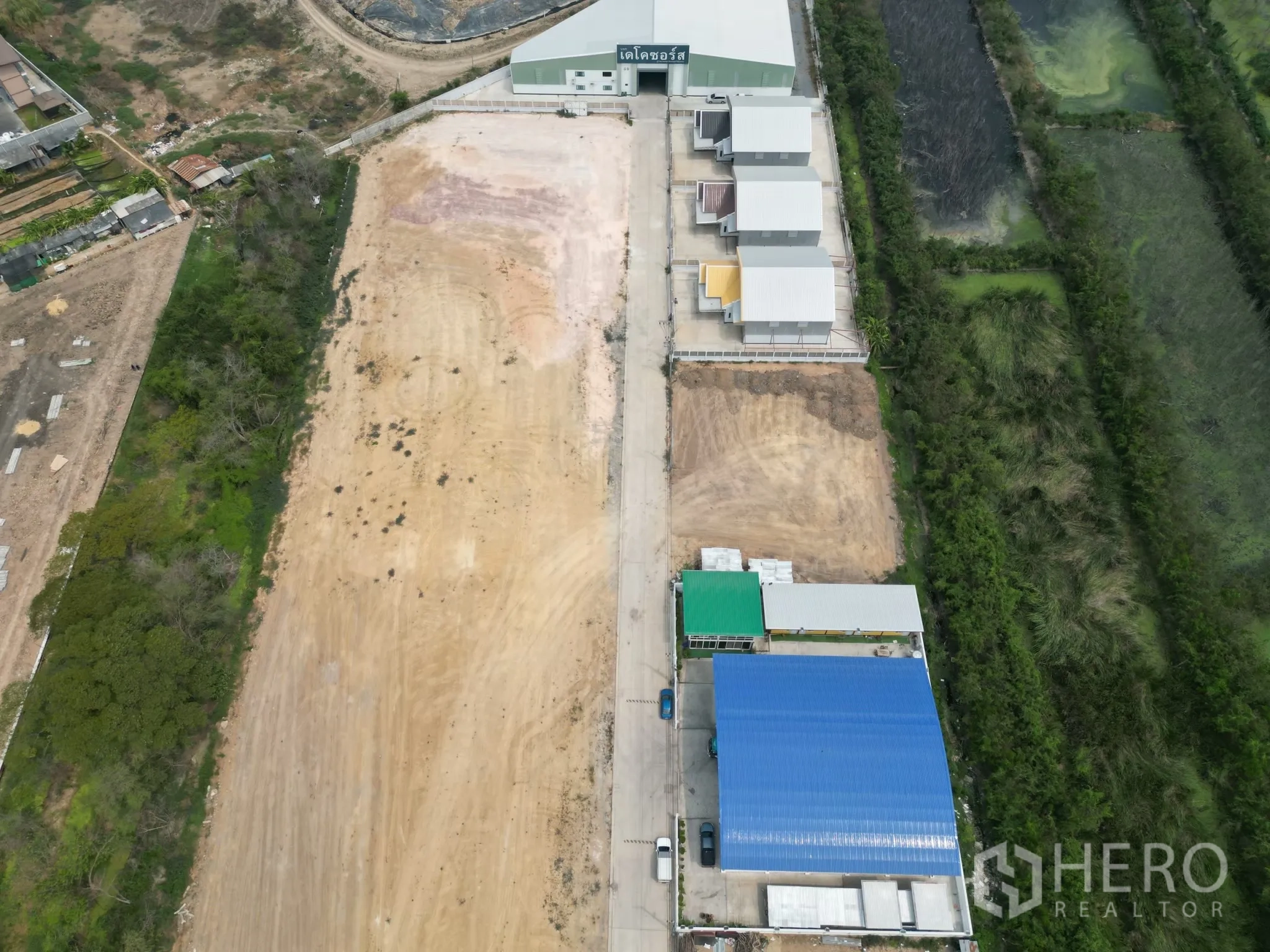Land for sale in Sai Noi, Nonthaburi - Aerial view of a large cleared plot bordered by an access road, neighboring warehouses, and surrounding greenery and water features.