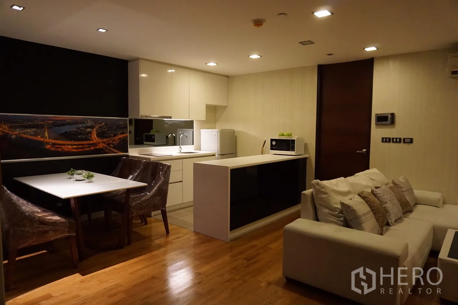 Condominium for rent or sale in Bang Rak, Bangkok - Open-plan living area in a Silom condo featuring a white modern kitchen with island, dining table, sofa and warm wooden flooring.