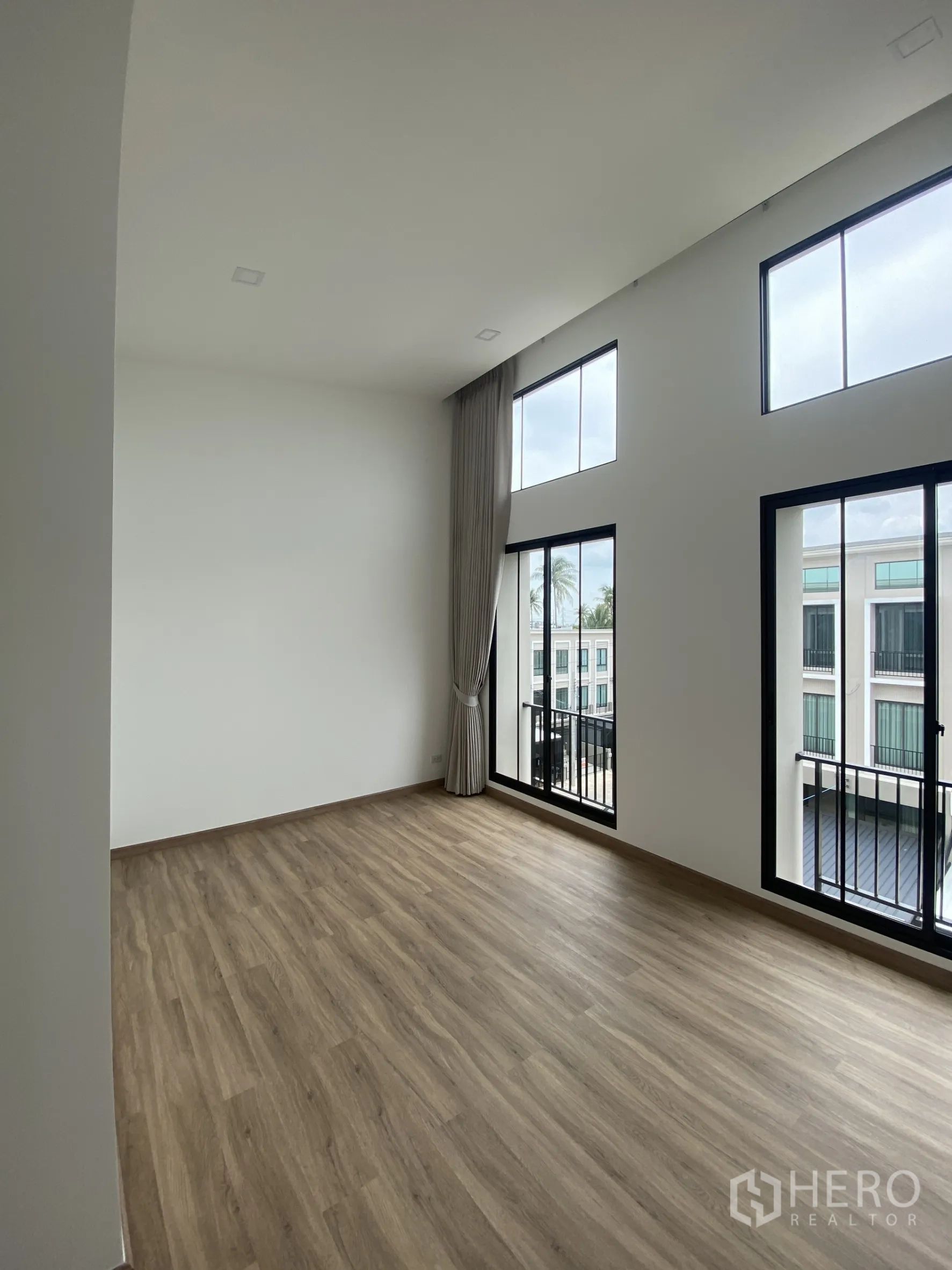Townhouse for rent or sale in Bang Khae, Bangkok - Sunlit room with high ceiling, wood floor, and wall of windows opening to Juliet balconies