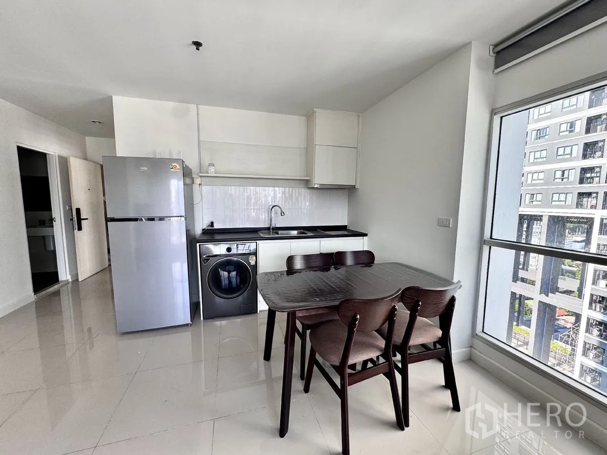 Condominium for rent in Huai Khwang, Bangkok - Kitchen and dining corner with fridge, sink, washer, and a four-seat table beside windows.