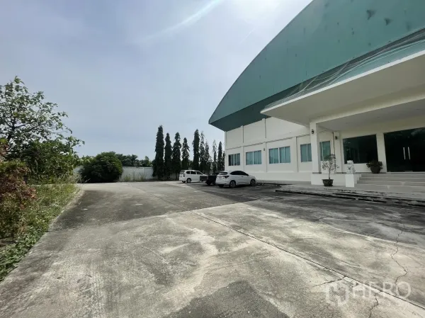 Warehouse for sale in Lat Lum Kaeo, Pathum Thani - Wide concrete driveway and the main entrance of the warehouse with landscaping and parked cars.