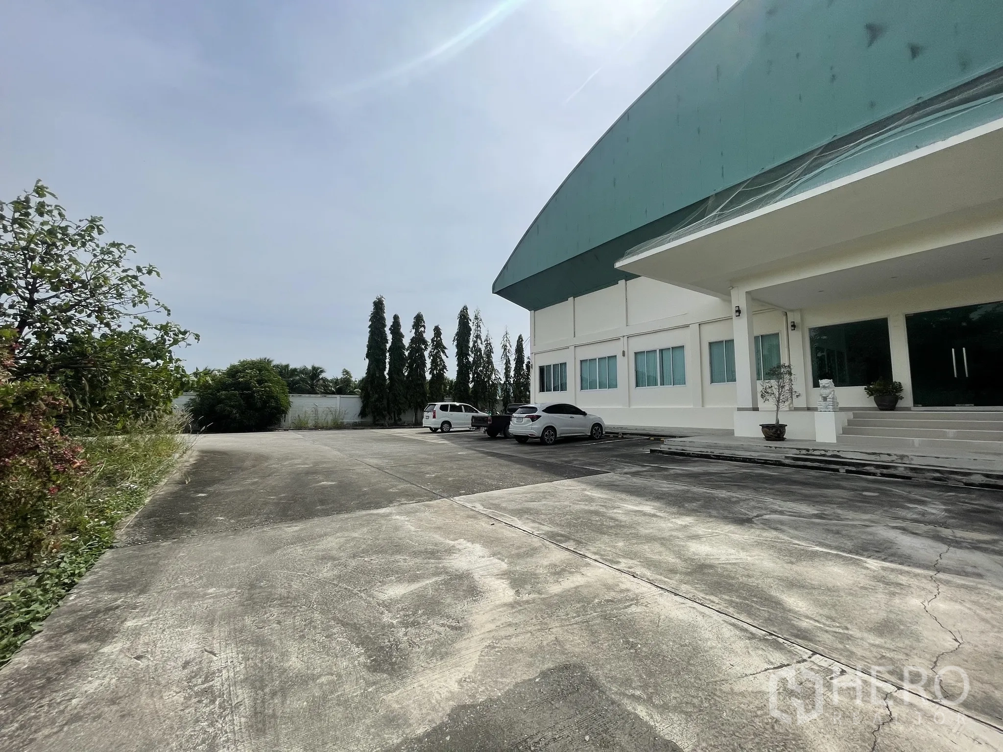 Warehouse for sale in Lat Lum Kaeo, Pathum Thani - Wide concrete driveway and the main entrance of the warehouse with landscaping and parked cars.