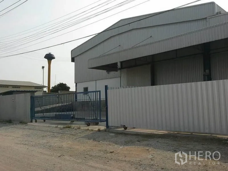 货栈 出租在Chon Buri - 仓库外立面与围栏滑动门，临宽阔道路，可见水塔