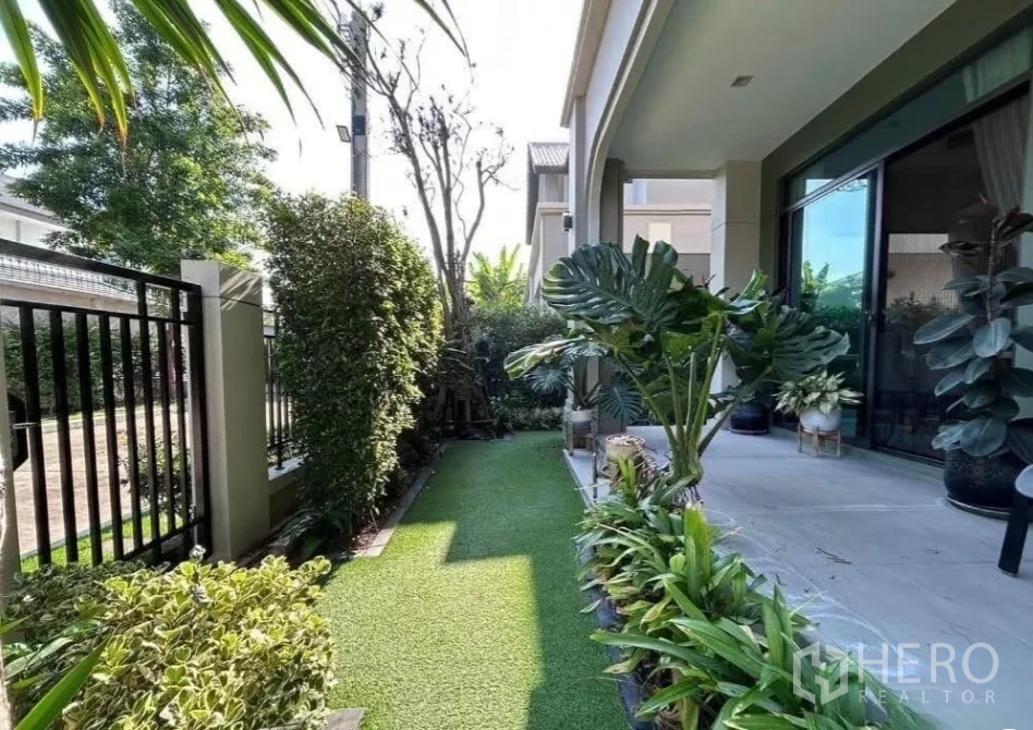 Detached House for rent in Bang Phli, Samut Prakan - Shaded side garden with artificial lawn, lush planters and a covered patio beside sliding glass doors.