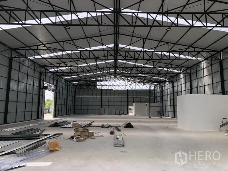 Warehouse for rent in Pak Kret, Nonthaburi - Open industrial hall with tall roof, metal walls and construction materials on the floor.