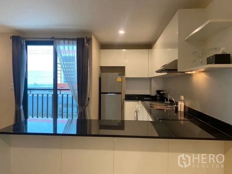 Condominium for rent in Watthana, Bangkok - Modern kitchen with black stone countertop, white cabinets and balcony door with sheer curtains.