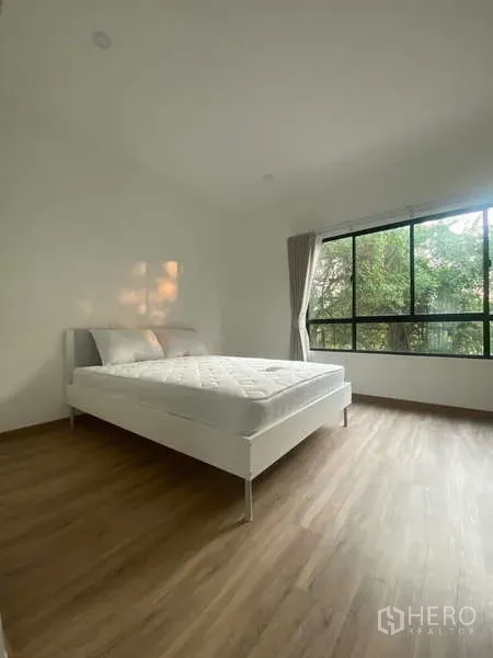 Townhouse for rent in Bang Kapi, Bangkok - Bedroom with wide black-framed windows facing greenery, minimal furniture and wood floors.