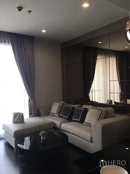 Condominium for rent in Watthana, Bangkok - Modern living room with L-shaped sofa, floor-to-ceiling curtains, mirrored wall and round black coffee table.