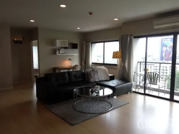 Condominium for rent or sale in Pathum Wan, Bangkok - Bright living area with a black leather sectional sofa, round glass coffee table, and balcony doors with city views.