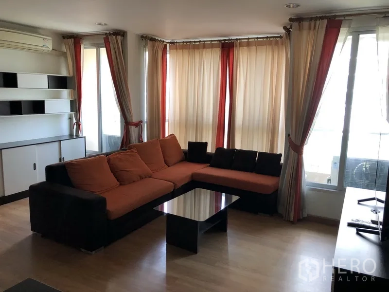 Condominium for rent in Khlong Toei, Bangkok - Bright living room with L-shaped sofa, full-height windows, sheer curtains, and balcony access.