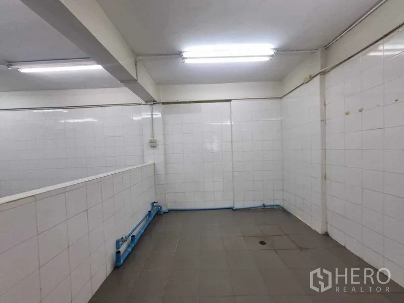 零售空间 出租在Bang Rak, Bangkok - 后场白色瓷砖房，荧光灯与蓝色管线，适合清洗或备餐。