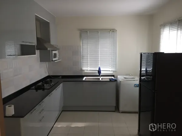 Detached House for rent in Pathum Thani - L-shaped kitchen with black countertops, gas hob, extractor hood, fridge and washing machine.