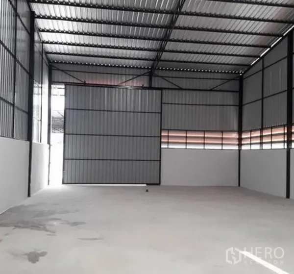 Warehouse for rent in Lam Luk Ka, Pathum Thani - Spacious warehouse with a tall metal sliding door partly open, corrugated steel walls and concrete floor.