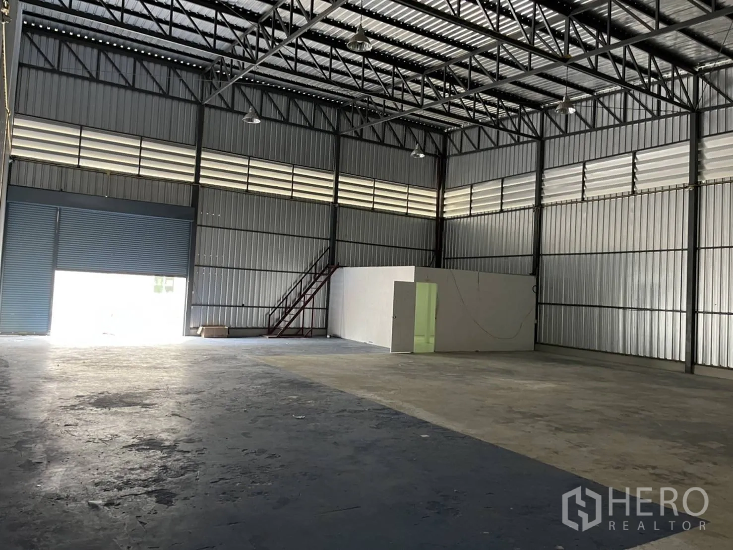 Warehouse for rent in Bang Phli, Samut Prakan - Empty warehouse hall with skylights, a roller shutter, and a metal stair beside the office block.