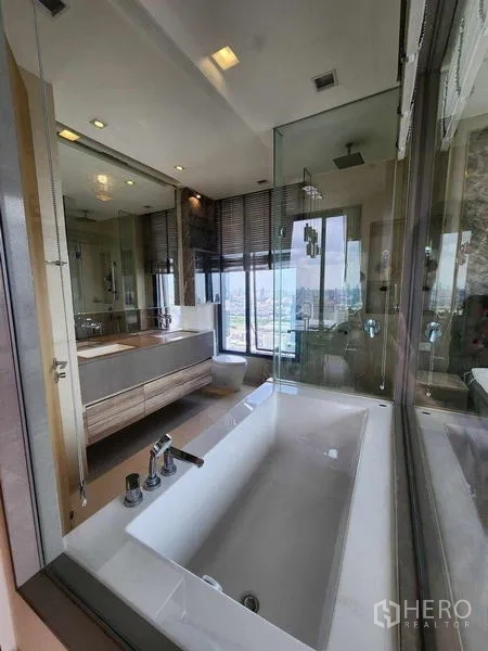 คอนโด ให้เช่า - ภาพใกล้อ่างอาบน้ำข้างผนังกระจก พร้อมเคาน์เตอร์อ่างล้างหน้าและวิวเมือง