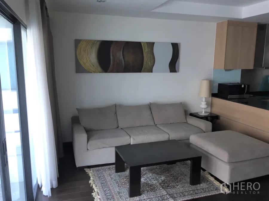 公寓 出租在Sathon, Bangkok - 采光良好的客厅，米色沙发、抽象装饰画、茶几与通往阳台的推拉门。