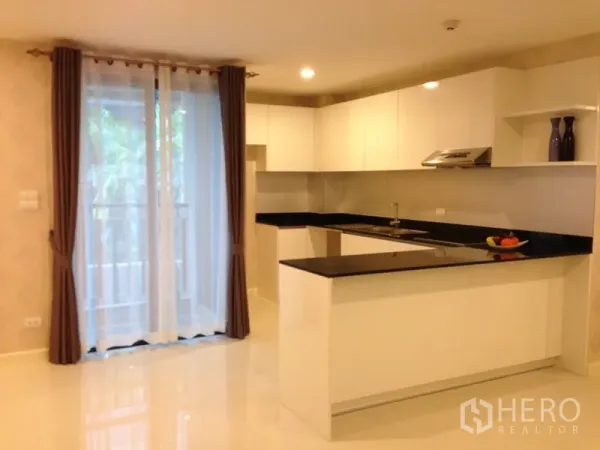 Condominium for rent or sale in Watthana, Bangkok - Open‑plan white kitchen with black stone countertops and sliding door to a balcony in a Bangkok condominium.