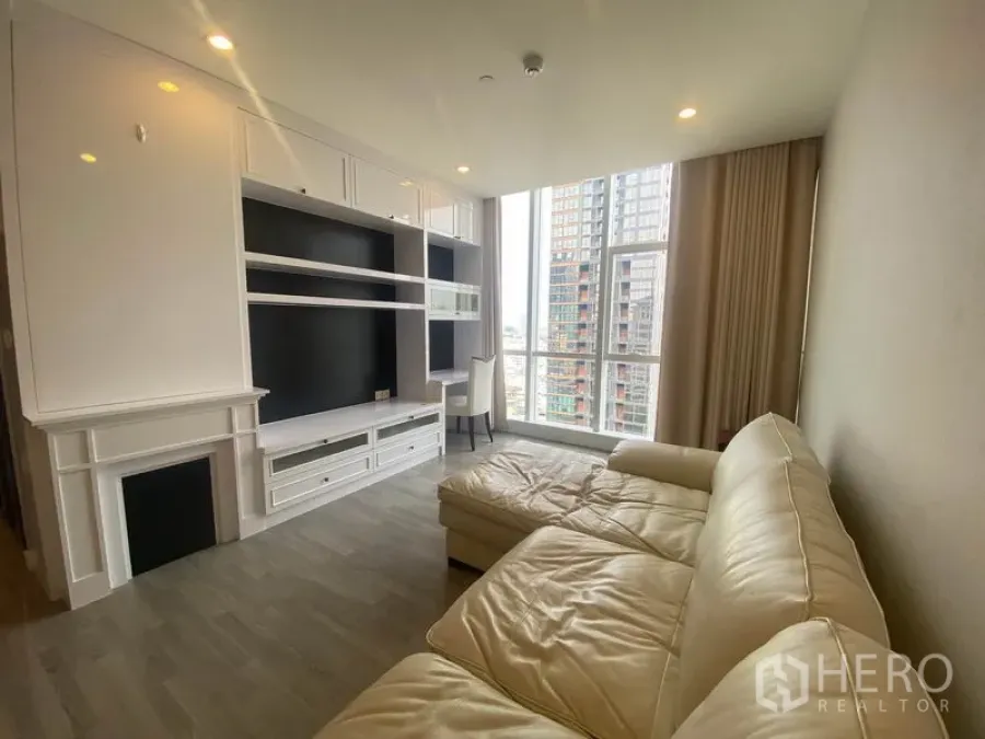 公寓 出租在Bang Rak, Bangkok - 明亮客厅，白色定制电视墙与L形沙发，落地窗俯瞰曼谷天际线。