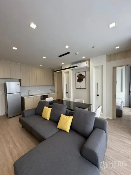 Condominium for rent in Watthana, Bangkok - Open-plan living room with a gray sectional, yellow cushions, light-wood kitchen and stainless refrigerator.