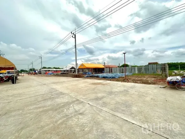 Land for sale in Lam Luk Ka, Pathum Thani - Cleared land plots with boundary fencing, temporary structures and power lines along the project road.