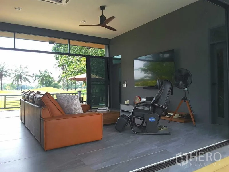 บ้าน ให้เช่าหรือขายในLam Luk Ka, Pathum Thani - ห้องนั่งเล่นพร้อมโซฟาหนัง พัดลมเพดาน ทีวีจอใหญ่ และประตูกระจกสไลด์วิวแฟร์เวย์