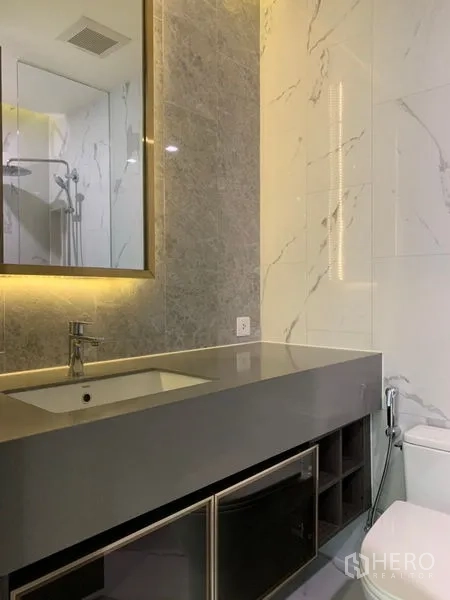 Condominium for sale in Khlong Toei, Bangkok - Secondary bathroom with long countertop, illuminated mirror and toilet.