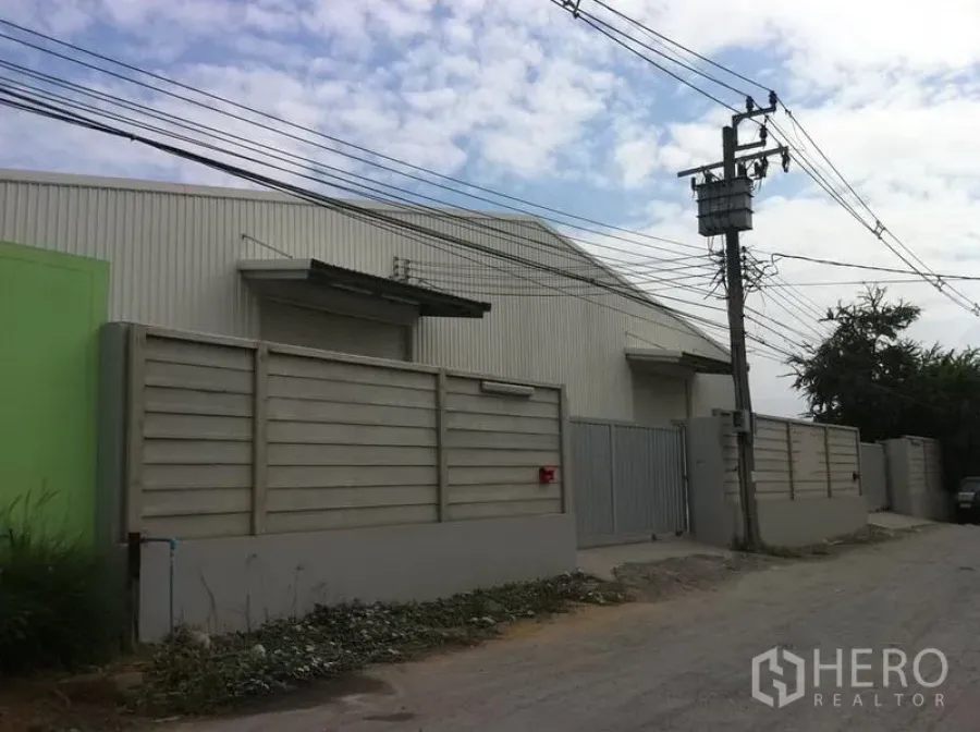 คลังสินค้า ให้เช่าในSai Mai, Bangkok - มุมมองจากถนนของโกดังรั้วรอบขอบชิด พร้อมประตูเหล็กทางเข้าและสายไฟเหนือศีรษะ ในเขตสายไหม กรุงเทพฯ