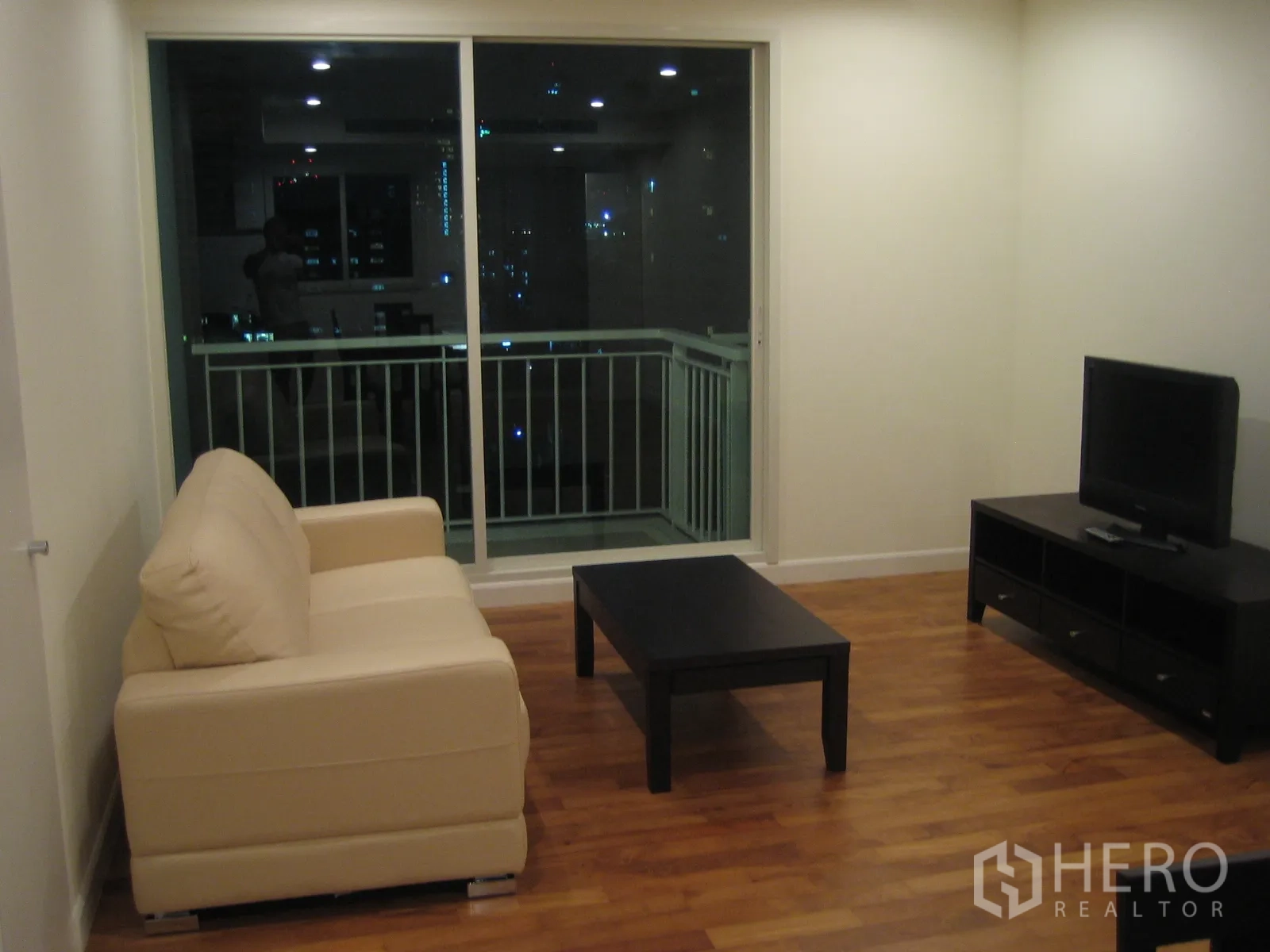Condominium for rent or sale in Watthana, Bangkok - Bright living room with beige sofa, TV console, wood flooring, and sliding glass doors to the balcony at night.