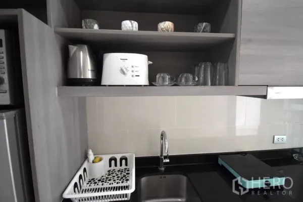 Condominium for rent in Khlong Toei, Bangkok - Compact kitchen with open shelves, kettle, toaster, glassware, single sink and black countertop.