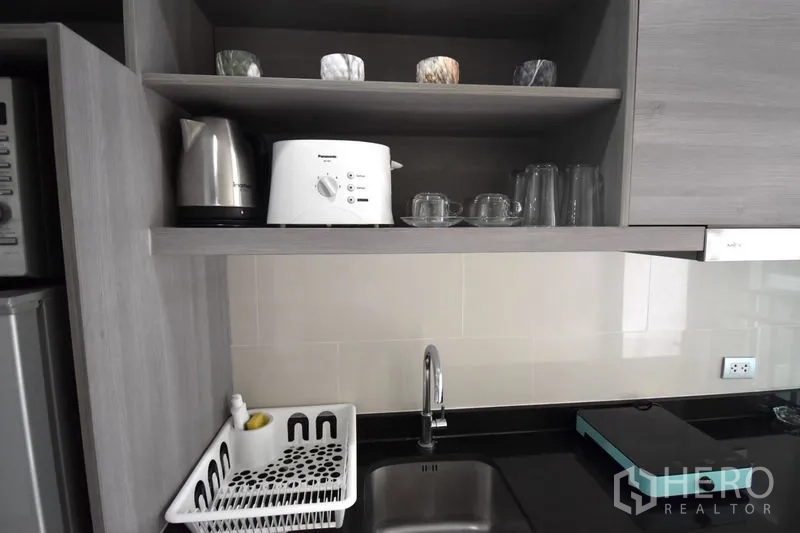 Condominium for rent in Khlong Toei, Bangkok - Compact kitchen with open shelves, kettle, toaster, glassware, single sink and black countertop.