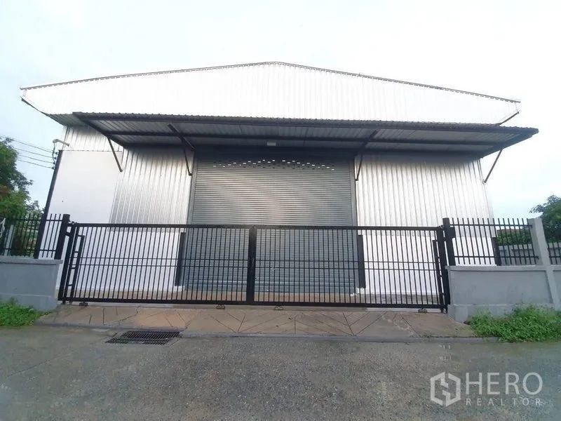 货栈 出租在Bang Khen, Bangkok - 仓库正面，宽幅卷闸门、遮雨棚与黑色金属围栏。