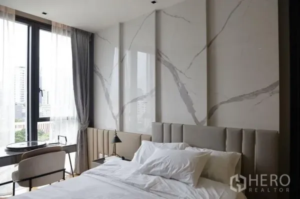 Condominium for rent in Khlong Toei, Bangkok - Bright bedroom with marble accent wall, floor-to-ceiling windows, city view, and a compact work desk and chair.