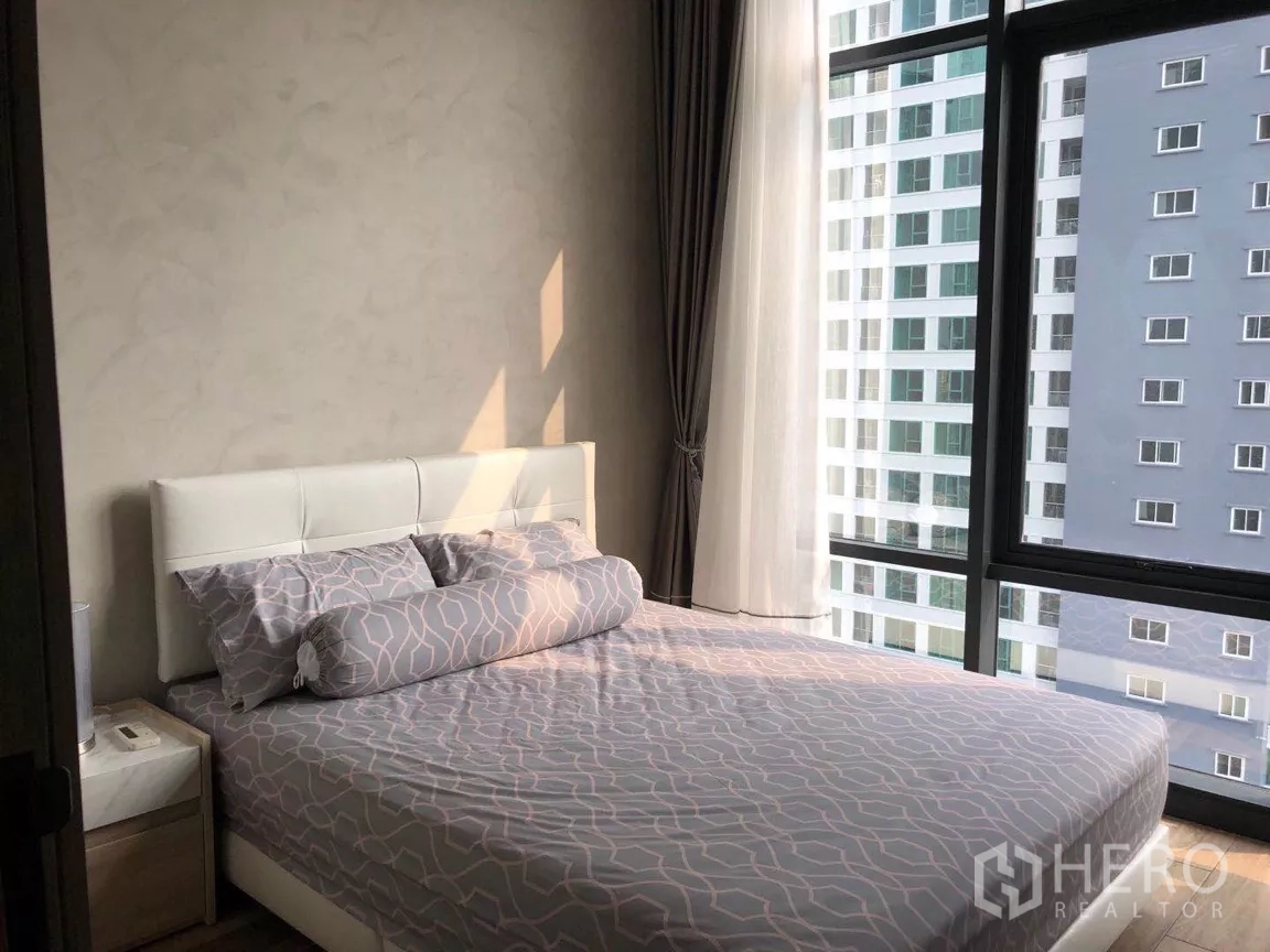 คอนโด ขายในWatthana, Bangkok - ห้องนอนรองเตียงสีขาว ชุดเครื่องนอนลายกราฟิก และหน้าต่างมุมห้องที่มองไปยังตึกข้างเคียง