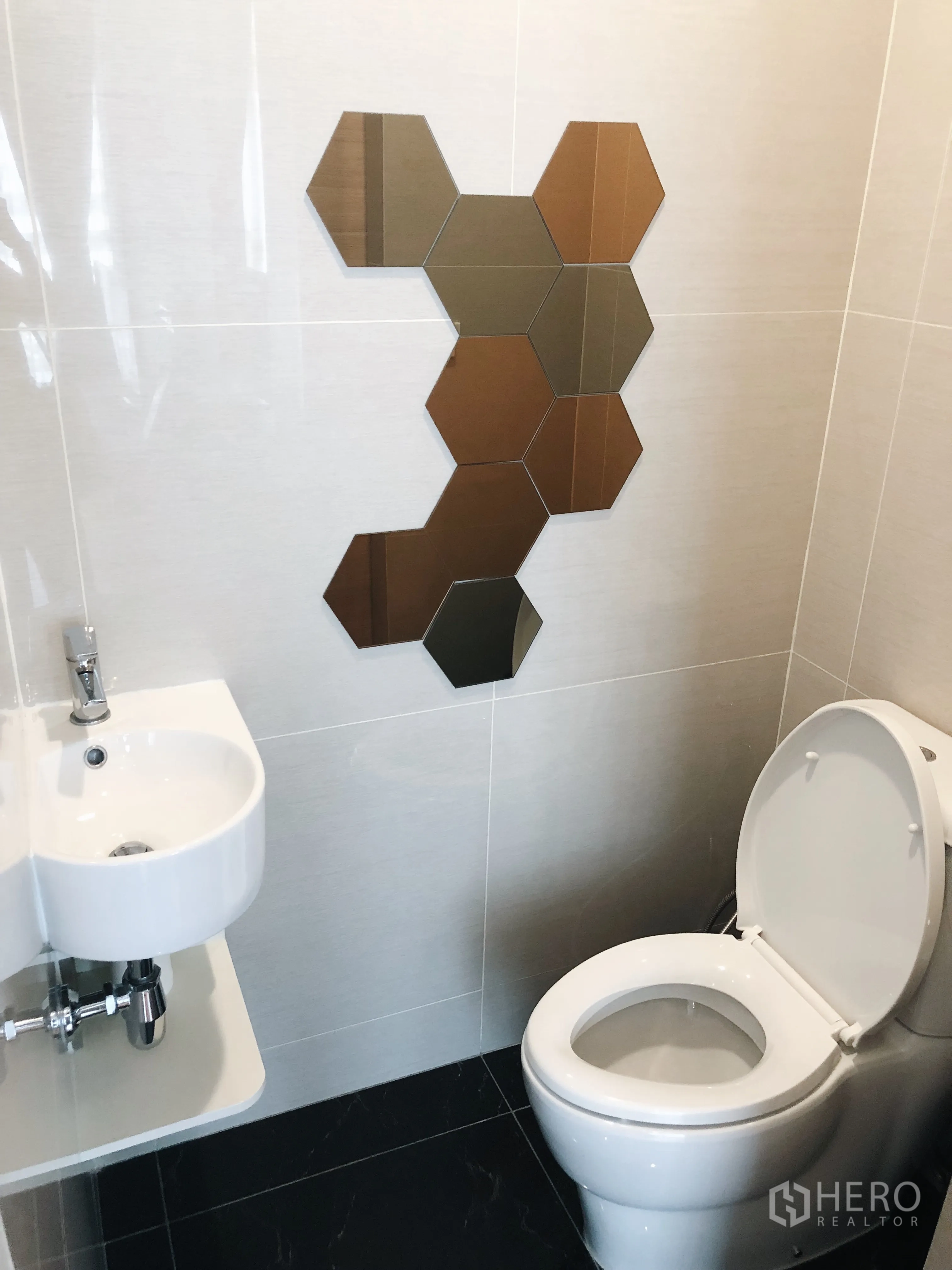 Condominium for rent in Khlong Toei, Bangkok - Contemporary bathroom with wall-mounted sink, toilet, and decorative hexagon mirror tiles on light walls.