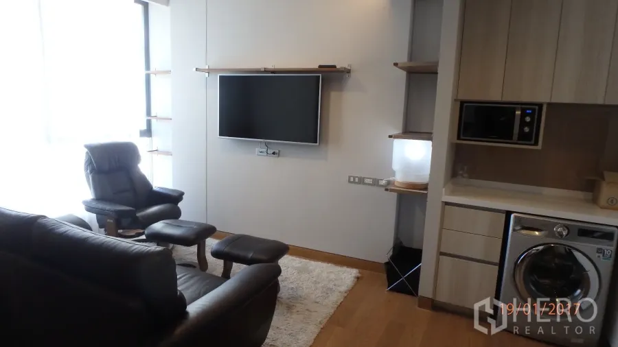 คอนโด ให้เช่าในKhlong Toei, Bangkok - ห้องนั่งเล่นสว่างเฟอร์นิเจอร์หนัง ทีวีติดผนัง หน้าต่างบานใหญ่ พื้นไม้ และแพนทรีพร้อมเครื่องซักผ้า