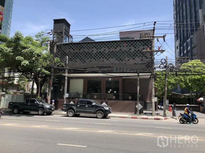 零售空间 出租在Watthana, Bangkok - 通罗临街的低层零售建筑，宽阔玻璃立面与抬高门廊，周边高楼与绿树，位于曼谷瓦他那区主干道。
