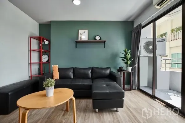 Condominium for rent in Watthana, Bangkok - Living room with black L-shaped sofa, green feature wall, and balcony access via sliding doors.