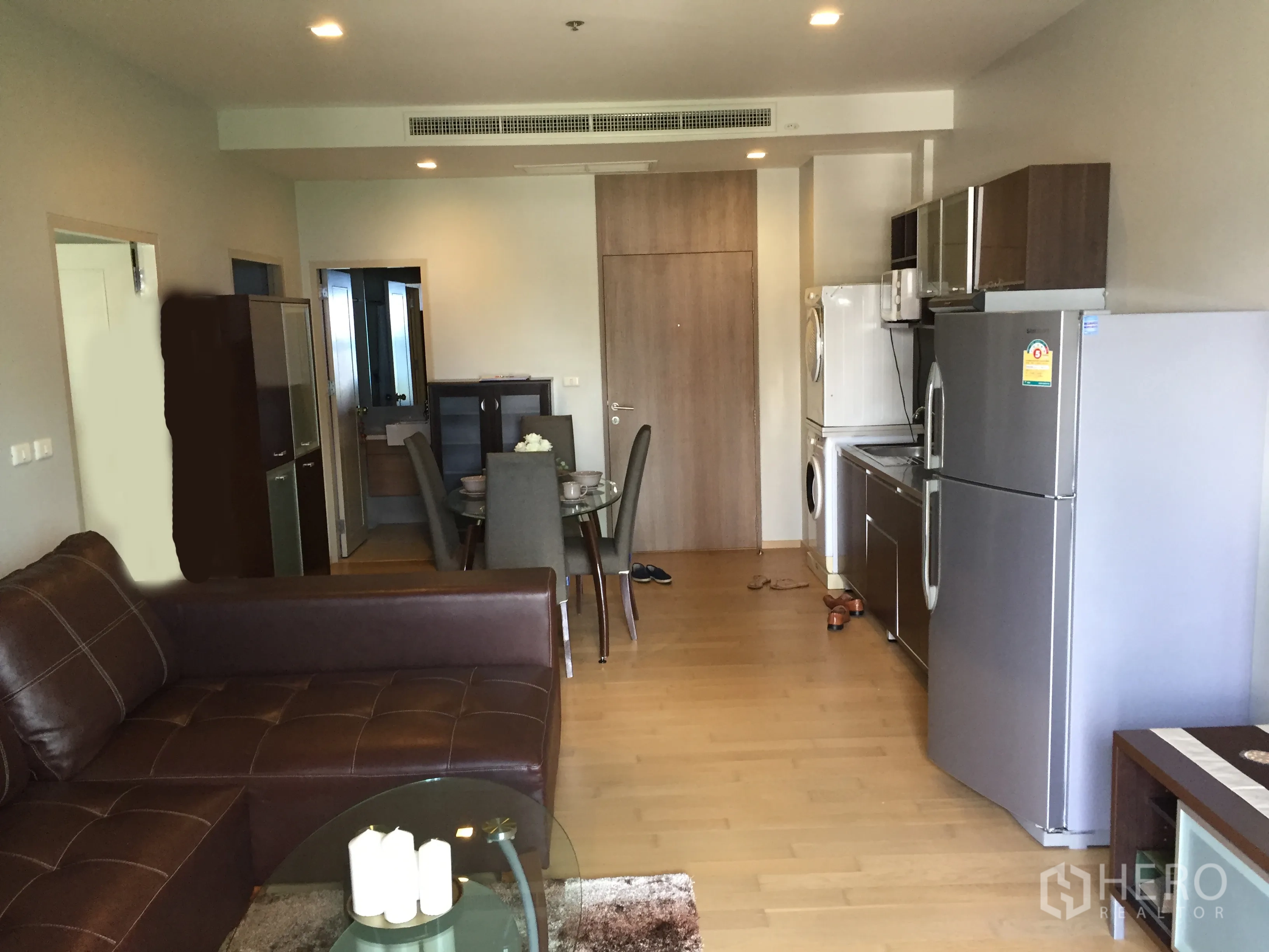 Condominium for rent in Watthana, Bangkok - Open‑plan living and dining area with leather sofa, wood floors, and fitted kitchen with appliances.