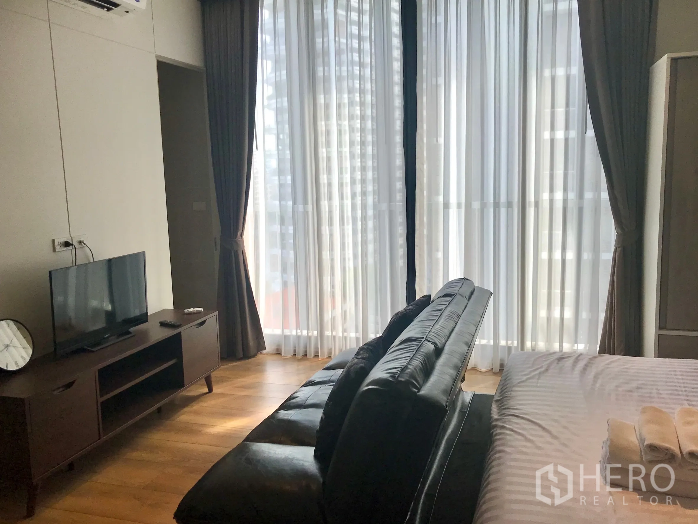 Condominium for sale in Khlong Toei, Bangkok - TV stand facing a sofa beside full-height windows with sheer curtains.