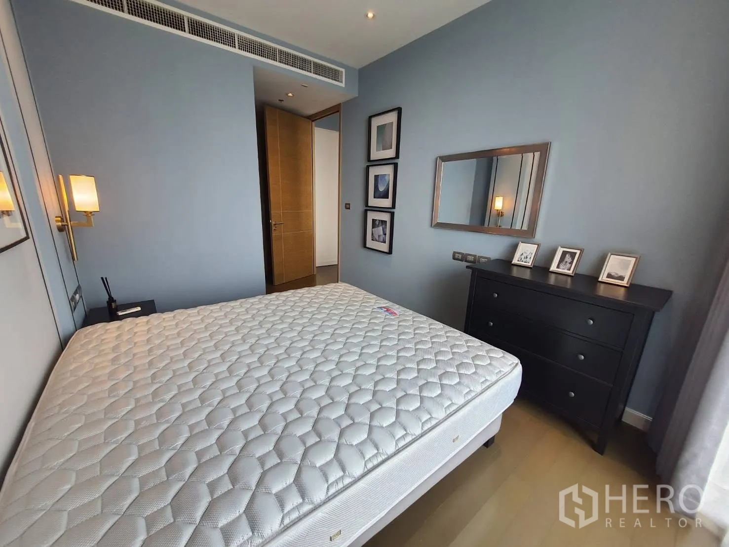 คอนโด for rent in Pathum Wan, Bangkok - ห้องนอนสว่าง ผนังสีฟ้า โคมไฟติดผนัง กระจกบานใหญ่ และตู้ลิ้นชักสีดำข้างเตียง