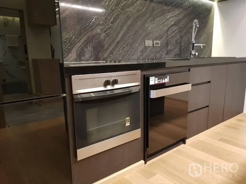 Condominium for rent in Bang Rak, Bangkok - Built-in oven and compact dishwasher beneath a stone countertop with textured marble backsplash.