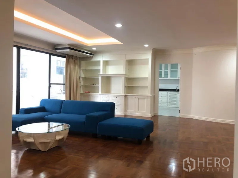 Condominium for rent in Watthana, Bangkok - Spacious living room with blue sectional sofa, built-in white shelving, parquet floors and large sliding windows.