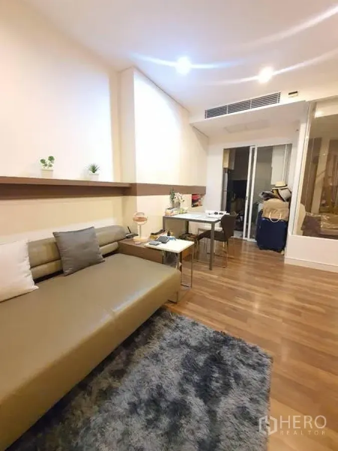 Condominium for sale in Bangkok - Living area with sectional sofa, textured rug, small dining table, and a glass partition to the bedroom.