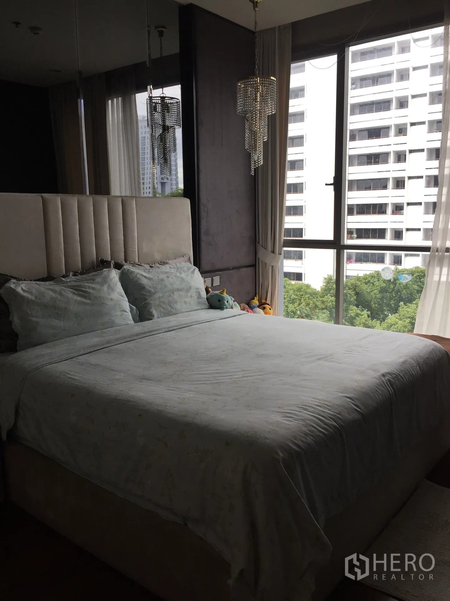 Condominium for sale in Watthana, Bangkok - Master bedroom with upholstered headboard, crystal pendant lights, and corner windows facing greenery and towers.