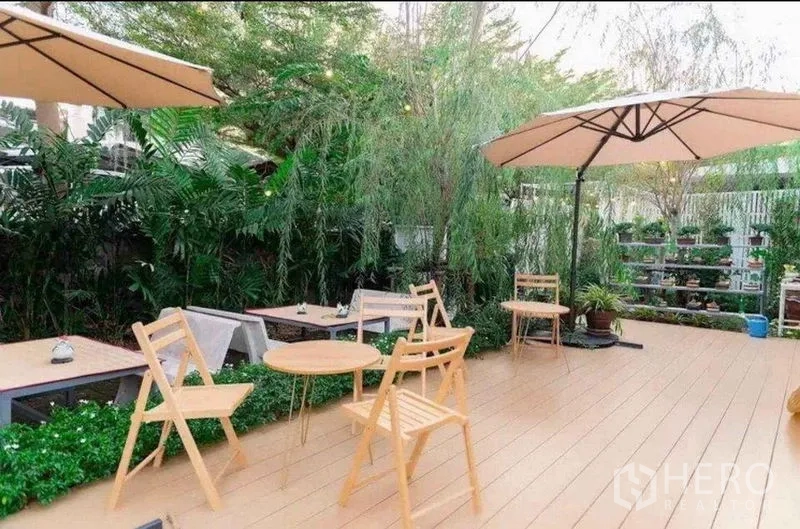 พื้นที่ขายปลีก ให้เช่าในSuan Luang, Bangkok - ระเบียงไม้กว้าง เก้าอี้และโต๊ะไม้โทนอ่อน ร่มสนาม และแนวต้นไม้ให้ความเป็นส่วนตัว