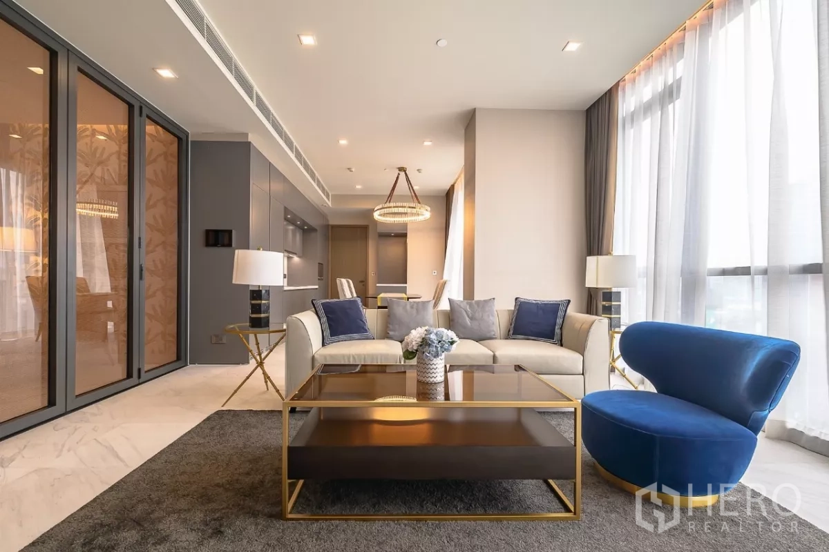 公寓 出租在Watthana, Bangkok - 现代客厅，米色沙发、蓝色单椅、玻璃茶几与一体式厨房。