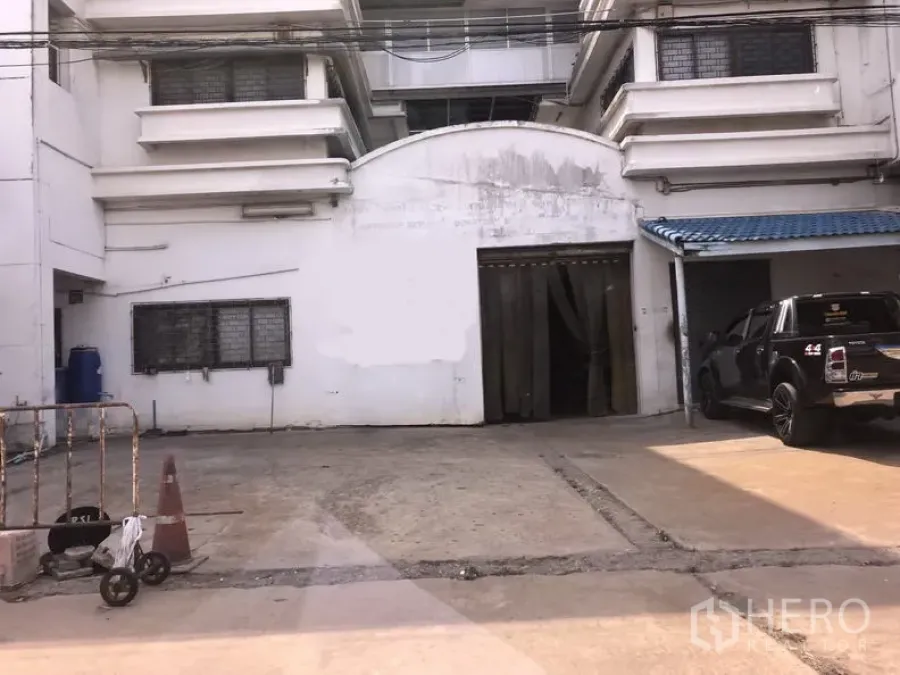 โรงงาน ขายในLam Luk Ka, Pathum Thani - ด้านหน้าโรงงานสีขาวที่ลำลูกกา มีลานคอนกรีต ประตูบานม้วนกว้าง และกันสีน้ำเงินพร้อมที่จอดรถข้างรถกระบะสีดำ